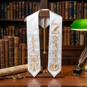 May include: A white graduation sash with gold embroidery, displayed on a wooden stand. The sash features the year "2024", along with decorative elements and text. The background includes a library setting with bookshelves and a desk.
