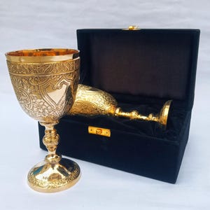 May include: A gold-colored metal chalice with intricate floral and heart-shaped engravings, featuring the word Mischief on its base. A second matching chalice rests inside a black velvet-lined presentation box with a gold-colored metal latch.