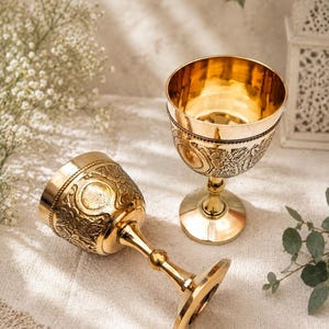 May include: Two polished gold-toned metal goblets featuring intricate embossed floral and circular patterns on the exterior bowls. Each goblet has a slender stem and a wide, circular base, with one standing upright and the other lying on its side.