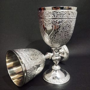 Puede incluir: Dos cálices de metal color plata con intrincados patrones florales y de volutas en relieve en el exterior de las copas y las bases. Cada cáliz tiene un borde interior liso y pulido, y un tallo decorativo delgado que conecta con una base ancha.