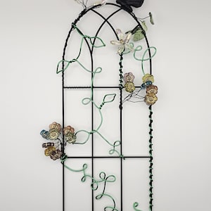 May include: A decorative black metal trellis with a butterfly and floral accents. The trellis features a black butterfly at the top, with green wire vines and colorful beaded flowers. The trellis is designed to be mounted on a wall.