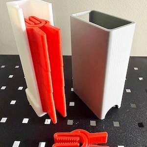 May include: A set of three-dimensional, geometric objects. A white rectangular container holds three bright red, U-shaped clips. A gray rectangular container sits next to the white container. A single red clip rests on the surface.