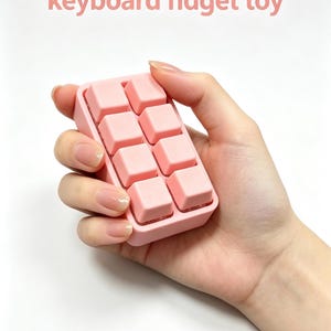 May include: A pink keyboard fidget toy with eight square keys. The toy is held in a hand against a white background. The text "keyboard fidget toy" is above the toy in pink.