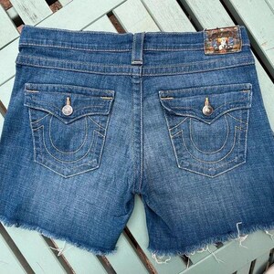 May include: Medium-wash blue denim shorts featuring a frayed raw hem, two back pockets with button-down flaps, and a rectangular leather-style brand patch on the back waistband. The pockets are detailed with thick, gold-toned contrast stitching.