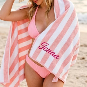May include: A pink and white striped beach towel with the name "Jenna" embroidered in a cursive font. The towel is wrapped around a person wearing a pink bikini and a straw hat. The beach scene includes the ocean and sand.