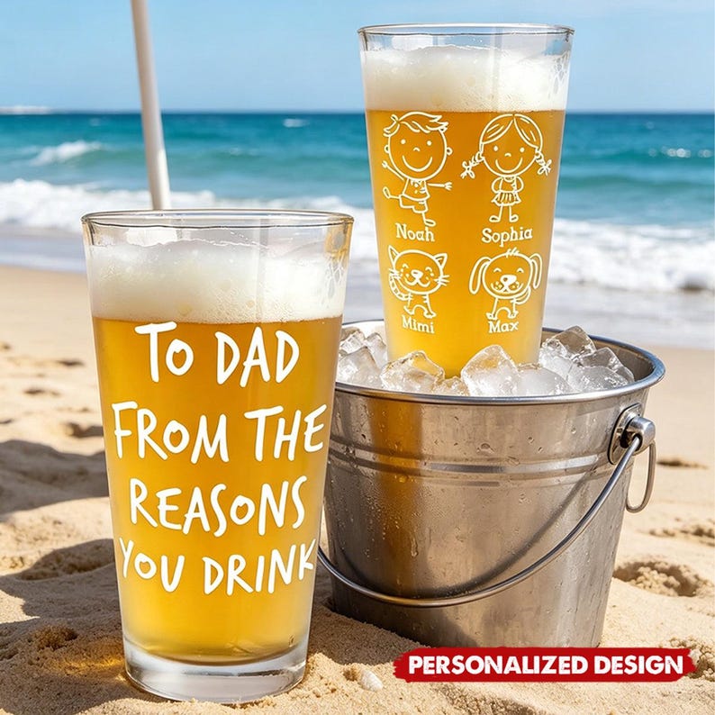 May include: Two clear pint glasses filled with beer, one with the text "TO DAD FROM THE REASONS YOU DRINK." The other glass has cartoon drawings of a boy, girl, cat, and dog with names. The glasses sit on a sandy beach next to a metal bucket filled with ice.
