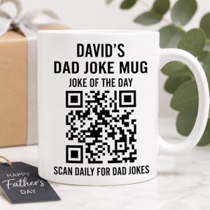 May include: A white ceramic mug featuring black text that reads DAVID'S DAD JOKE MUG and JOKE OF THE DAY above a large black and white QR code, with the text SCAN DAILY FOR DAD JOKES printed below the code, accompanied by a black gift tag.