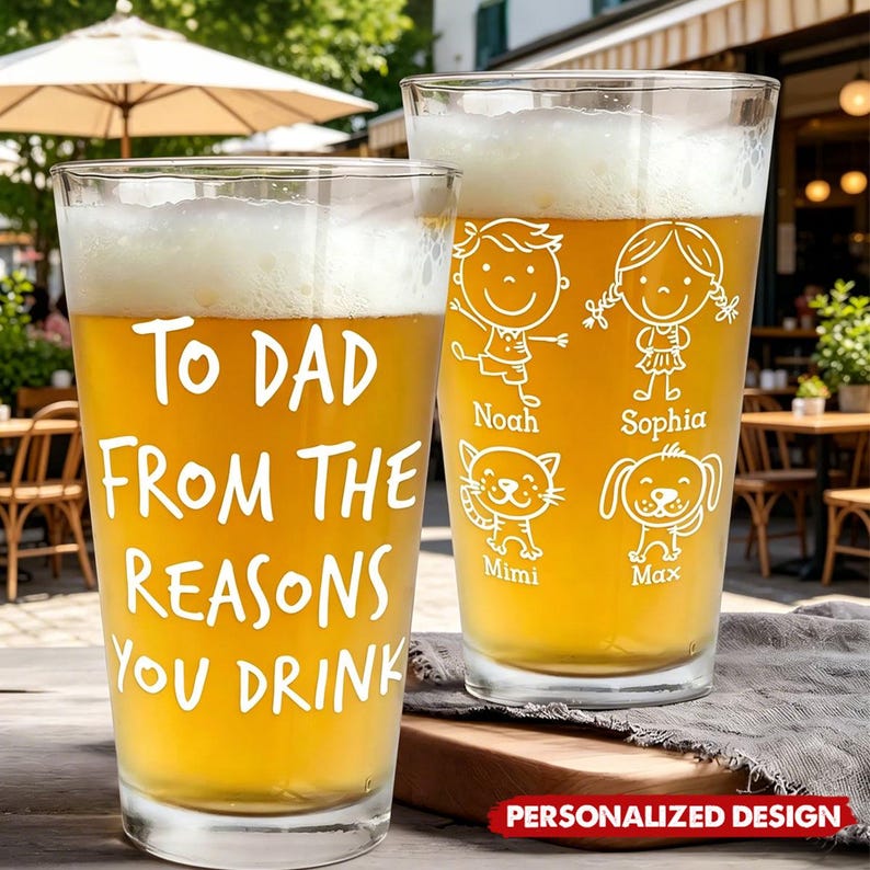 May include: Two clear pint glasses filled with beer. One glass has the text "TO DAD FROM THE REASONS YOU DRINK". The other glass has cartoon drawings of a boy, girl, cat, and dog with names. The glasses are on a wooden surface.