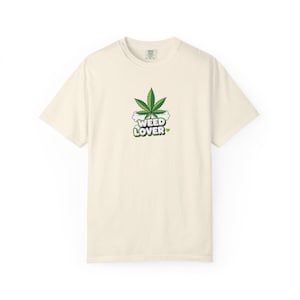 Puede incluir: Camiseta color crema con un diseño de hoja de cannabis verde y las palabras "WEED LOVER". El diseño incluye un hueso y un corazón. La camiseta tiene mangas cortas y cuello redondo.