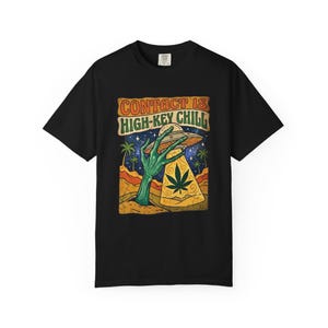 Puede incluir: Camiseta negra con un gráfico que presenta una mano alienígena verde sosteniendo un OVNI sobre un paisaje desértico. El OVNI tiene una hoja de cannabis. El texto en la camiseta dice "CONTACT IS HIGH-KEY CHILL."