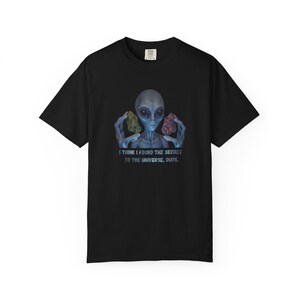 Puede incluir: Camiseta negra con un alienígena azul sosteniendo dos objetos coloridos. El alienígena tiene ojos grandes y está centrado en la camiseta. El texto debajo dice: "I THINK I FOUND THE SECRET TO THE UNIVERSE, DUDE."