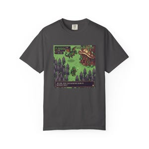 Puede incluir: Camiseta gris oscuro con un gráfico de videojuego pixelado. La escena del juego incluye un dragón, una casa de champiñones y un personaje con una antorcha. El juego se titula "Cannabis Quest" y el texto dice "¡Bienvenido Héroe! Tu aventura comienza en Weedwood Forest."