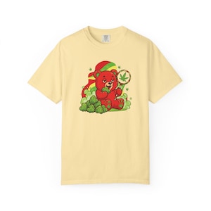 Puede incluir: Camiseta amarillo claro con un oso rojo de dibujos animados sosteniendo una hoja de cannabis y rodeado de cogollos de cannabis verdes. El oso lleva un sombrero rojo y amarillo. La camiseta es una prenda informal.