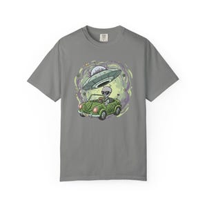 Puede incluir: Camiseta gris con una ilustración de dibujos animados de un extraterrestre conduciendo un coche descapotable verde con un perro, debajo de un OVNI. El diseño incluye un fondo verde y morado arremolinado, y el extraterrestre lleva gafas de sol.