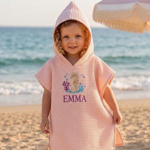 Può includere: Un telo da mare con cappuccio rosa chiaro con ricamo di cavalluccio marino e corallo. L'asciugamano ha il nome "EMMA" ricamato in viola. Il bambino indossa l'asciugamano su una spiaggia sabbiosa con l'oceano sullo sfondo.
