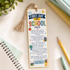 Könnte beinhalten: Ein rechteckiges Lesezeichen aus Papier mit einer Jutequaste und einer Holzperle. Der Text lautet First Day of School Prayer, gefolgt von einem Gebet und Illustrationen eines roten Apfels, eines Rucksacks, eines Globus, Büchern und Stiften.