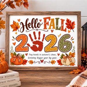 May include: A framed sign featuring the text Hello FALL 2026 with a red handprint replacing the zero. The design includes orange and green polka-dot numbers, autumn leaves, pumpkins with plaid bows, and the text Tiny hands in autumn's cheer, Growing bigger year by year.