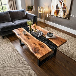 Puede incluir: Una mesa de centro rectangular con un tablero de madera natural de borde vivo y una incrustación central de resina negra. La mesa está sostenida por dos patas de metal negro en forma de U y muestra un patrón de veta de madera visible.