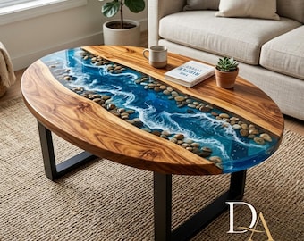 May include: Oval wooden coffee table with a river design. The center features a blue resin river with white wave details and embedded stones. The table has a dark wood frame and black metal legs. A book and a mug sit on the table.