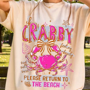 以下が含まれることがあります： ピンクのカニのグラフィックと「if Crabby feeling beachy PLEASE RETURN TO THE BEACH」という文字が入った、クリーム色の半袖シャツ。デザインには、リボン、ヒトデ、貝殻が含まれています。