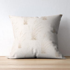 May include: A square throw pillow featuring a light beige fabric base with a repeating pattern of delicate, thin, tan-colored stalks of wild grass. The design is printed across the entire surface of the pillow, creating a soft, natural aesthetic.