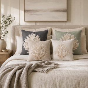 May include: A collection of square throw pillows arranged on a bed, featuring embroidered coral motifs in shades of cream and beige. The pillows are presented in charcoal gray, sage green, and neutral cream fabric covers, layered against a headboard.