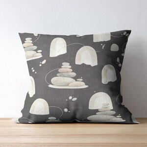 May include: A square throw pillow featuring a dark charcoal gray background with a repeating pattern of stacked smooth stones in shades of cream, beige, and light gray, interspersed with arched shapes and thin, curved white line accents.