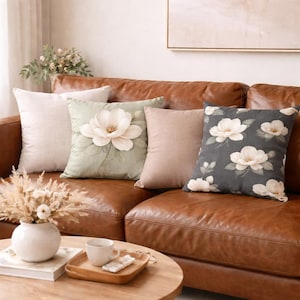 May include: A collection of decorative throw pillows on a brown leather sofa. The pillows feature various designs, including a large floral applique, solid colors, and a floral pattern on a dark background. The pillows add a touch of elegance to the living room.