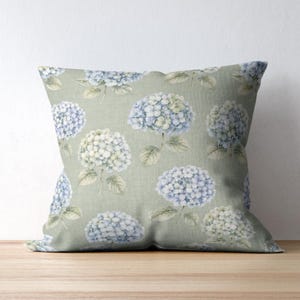 May include: Square throw pillow with a light green background and a repeating pattern of blue and white hydrangea flowers. The pillow is made of a soft fabric and is designed for decorative use.