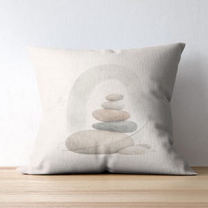 May include: A square throw pillow featuring a printed design of a stacked stone cairn in muted shades of beige, gray, and soft blue. The stones are arranged in a vertical pile centered against a faint, arched watercolor-style background graphic.