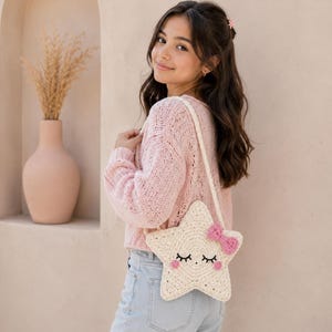 May include: A cream-colored crochet star-shaped crossbody bag featuring a pink bow, embroidered closed eyes with eyelashes, and pink circular cheeks. The bag has a long matching strap and is worn over a light pink knit sweater and light blue jeans.