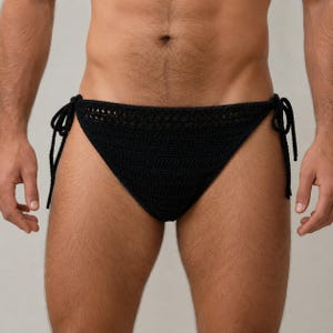 May include: A black crochet bikini bottom featuring a textured knit pattern and adjustable side ties. The garment has a triangular front panel with a decorative openwork border along the top edge and thin strings for securing at the hips.