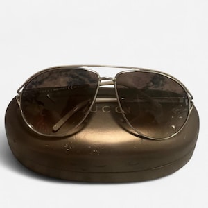 May include: Aviator-style sunglasses with a silver frame and gradient brown lenses. The sunglasses rest on a brown case. The brand name "GUCCI" is visible on the case. The lenses are oval-shaped.