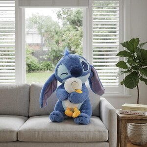 Puede incluir: Un gran peluche azul del personaje Stitch de Lilo & Stitch, abrazando a un pato de peluche blanco y amarillo m&aacute;s peque&ntilde;o. Los peluches est&aacute;n en un sof&aacute; gris frente a una ventana con persianas blancas.