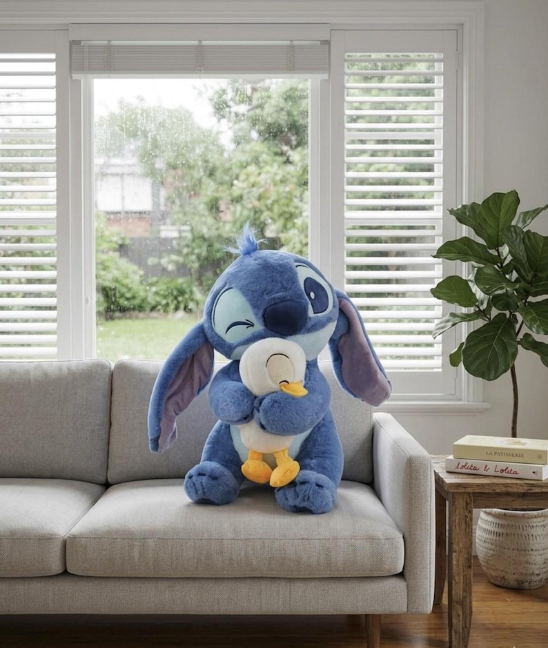 Puede incluir: Un gran peluche azul del personaje Stitch de Lilo & Stitch, abrazando a un pato de peluche blanco y amarillo m&aacute;s peque&ntilde;o. Los peluches est&aacute;n en un sof&aacute; gris frente a una ventana con persianas blancas.