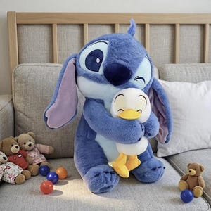 Puede incluir: Un gran peluche azul de Stitch abrazando a un peque&ntilde;o pato de peluche blanco y amarillo. El juguete Stitch tiene orejas grandes y una nariz azul oscuro. Varios peque&ntilde;os osos de peluche y bolas de colores est&aacute;n en el fondo.