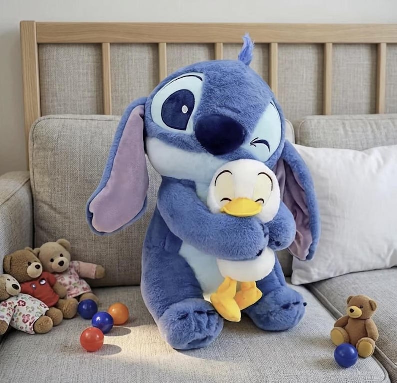 Puede incluir: Un gran peluche azul de Stitch abrazando a un peque&ntilde;o pato de peluche blanco y amarillo. El juguete Stitch tiene orejas grandes y una nariz azul oscuro. Varios peque&ntilde;os osos de peluche y bolas de colores est&aacute;n en el fondo.