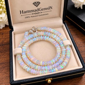 May include: A necklace made of faceted, round, translucent opal beads with iridescent flashes of color. The necklace is coiled inside a black jewelry box with a beige interior, featuring the text HammadGemsIN 100% Natural Gemstones.