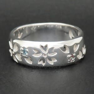 May include: A polished silver band ring featuring a repeating cutout pattern of five-petaled cherry blossom flowers. Two of the flower centers are accented with small, round, sparkling blue and pink gemstones set into the metal surface.