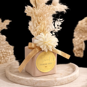 May include: A beige square vase with a gold ribbon and a gold circular tag that reads "Sweet. Thank You." The vase is filled with dried flowers and a white flower. The vase sits on a round, marble-effect tray.