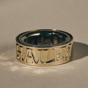 Könnte beinhalten: Ein goldfarbener Ring mit dem eingravierten Namen "HAILEY" um das Band. Der Ring hat eine polierte Oberfläche und ein dunkles Inneres. Die Zahl "5" ist ebenfalls auf dem Ring zu sehen.