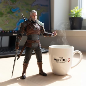 May include: A detailed figurine of Geralt of Rivia from The Witcher 3 video game, holding a sword. A white mug with "The Witcher 3 Wild Hunt" printed on it sits nearby, with steam rising. The figurine is in a detailed costume.