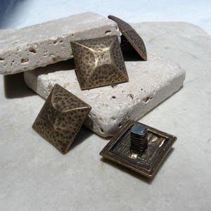 May include: Five antique brass pyramid-shaped tacks with a hammered finish. The tacks are arranged on a light beige stone surface.