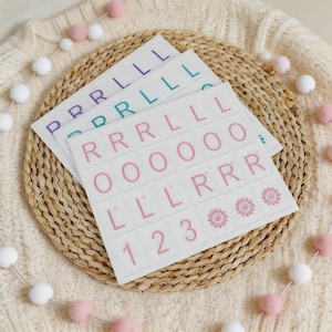 Könnte beinhalten: Drei Bögen weißer Aufkleber mit Buchstaben und Zahlen in Lila, Türkis und Rosa. Die Aufkleber sind auf einer gewebten Matte angeordnet, mit einer Girlande aus rosa und weißen Pompons im Hintergrund.