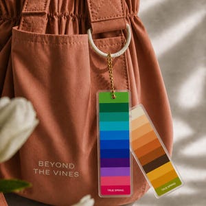 May include: A terracotta-coloured tote bag with a drawstring closure and a front pocket. The bag has a white ring and a gold chain holding two colour palette tags. The tags read "TRUE SPRING" and display colour gradients.