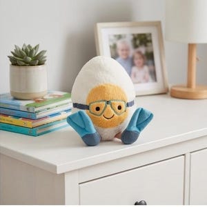 May include: A plush, egg-shaped toy with a yellow face, blue goggles, and blue flipper feet. The toy is white with a yellow face and a smiling expression. The toy is sitting on a white surface.