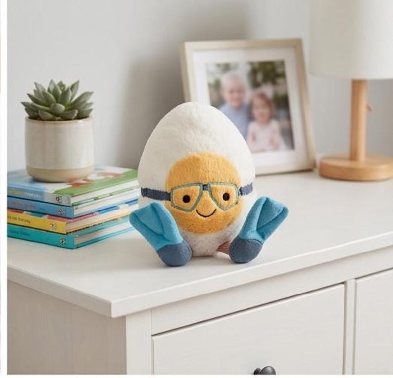 May include: A plush, egg-shaped toy with a yellow face, blue goggles, and blue flipper feet. The toy is white with a yellow face and a smiling expression. The toy is sitting on a white surface.