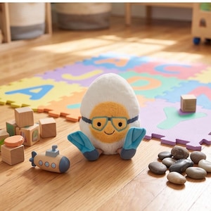 May include: A plush toy shaped like an egg with a smiling face, wearing blue flippers and goggles. A wooden submarine toy and building blocks are nearby. The background includes a colorful alphabet play mat and wooden floor.