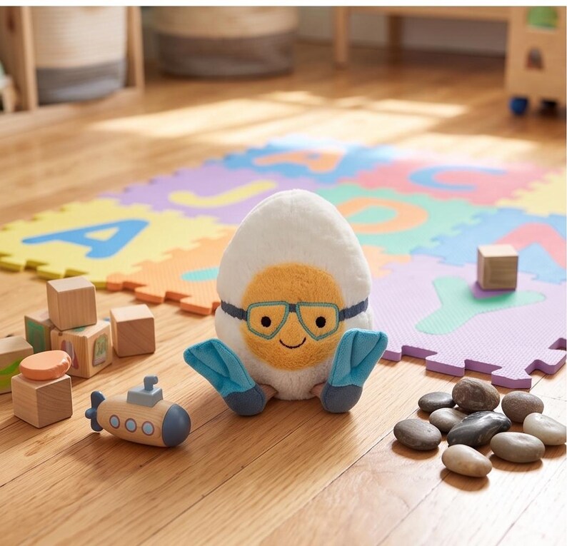 May include: A plush toy shaped like an egg with a smiling face, wearing blue flippers and goggles. A wooden submarine toy and building blocks are nearby. The background includes a colorful alphabet play mat and wooden floor.