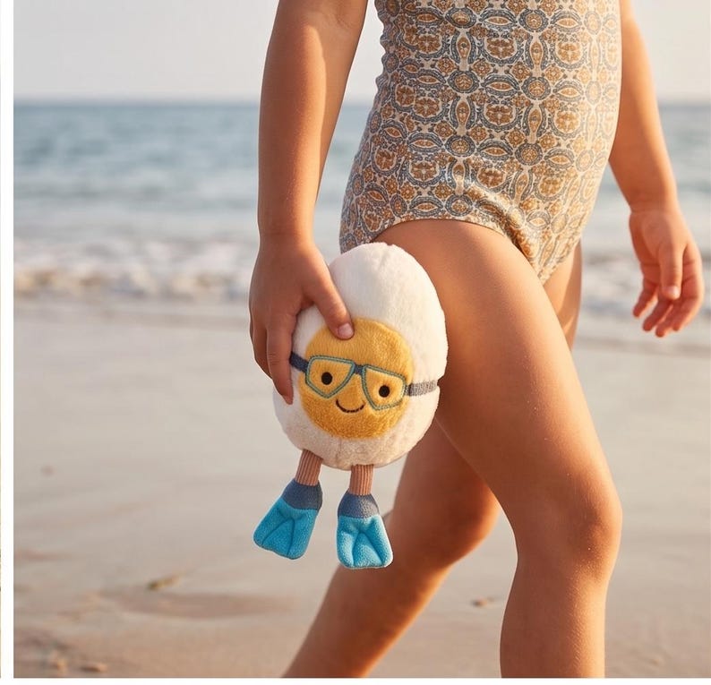 May include: A plush toy shaped like a sunny-side-up egg, wearing blue-tinted goggles and blue flipper feet. The egg has a smiling face with embroidered features. The toy is held by a person on a sandy beach, suggesting a summer or beach theme.